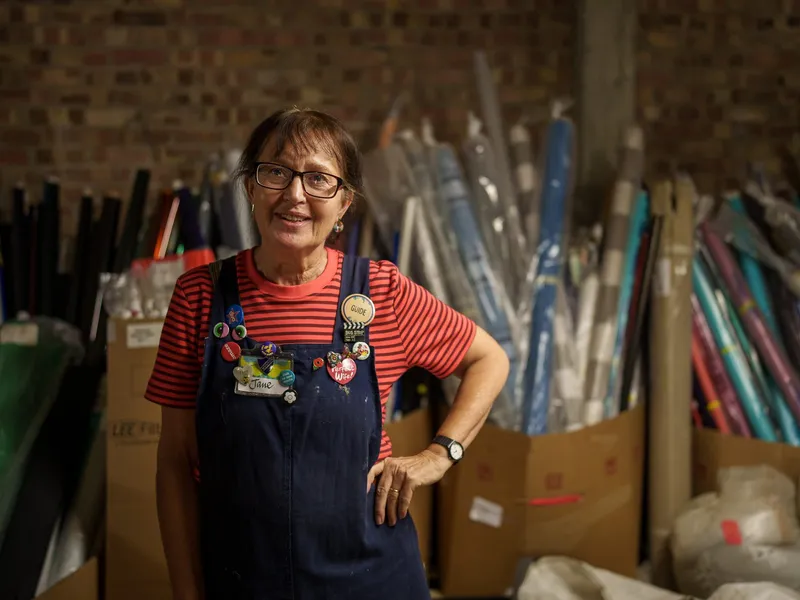 A Scrapstore volunteer in badge-covered dungarees, standing among rolls of paper and card
